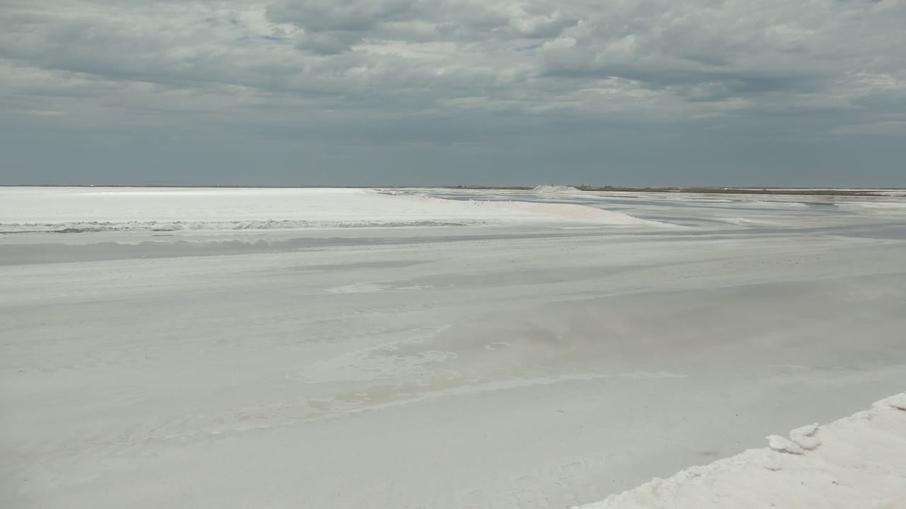 A pale ice and snow expanse in Guerrero Negro fills the foreground while a thin dark shoreline sits below layered gray clouds, with no visible people.