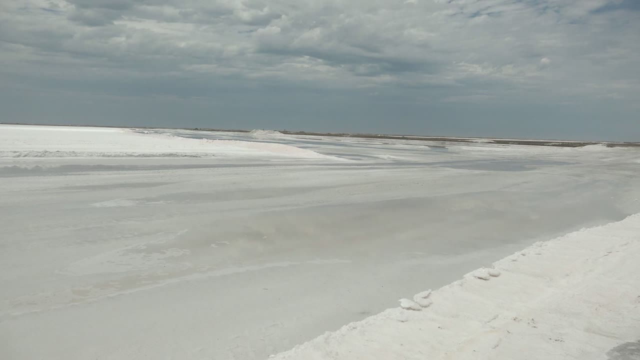 Wide ground-level view of a frozen waterway and white snow banks in Guerrero Negro, Baja California Sur, Mexico, with a dark overcast sky and no visible people.