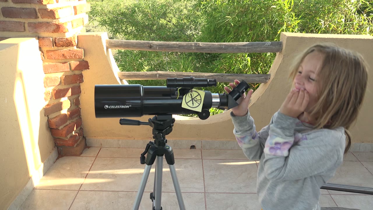 In San Carlos, Mexico, a child in a gray sweatshirt smiles with a hand near their mouth beside a black tripod-mounted telescope on a tiled patio.