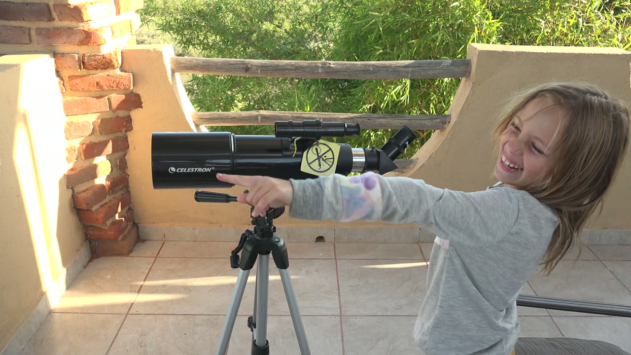 In San Carlos, Mexico, the child points toward the side of the black telescope while smiling, with the tripod centered on the tiled patio.