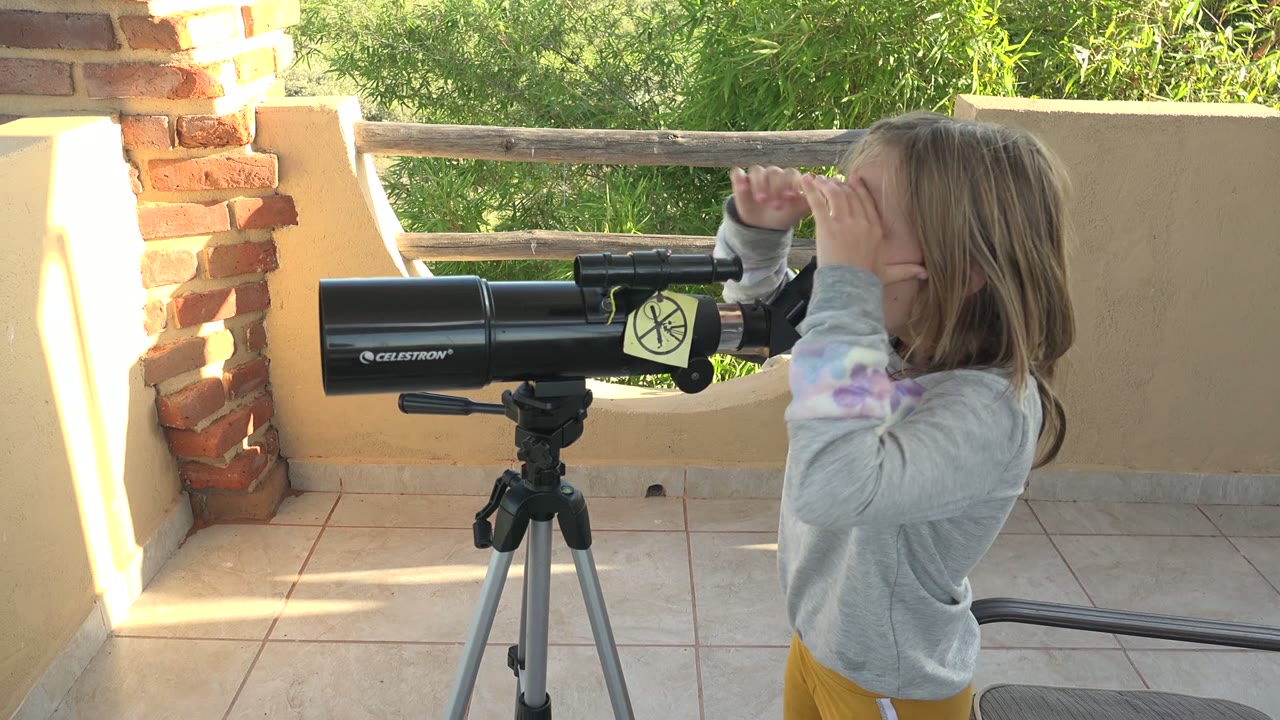 A young girl in San Carlos, Mexico looks through a black telescope mounted on a tripod beside brick columns and a tiled patio wall.