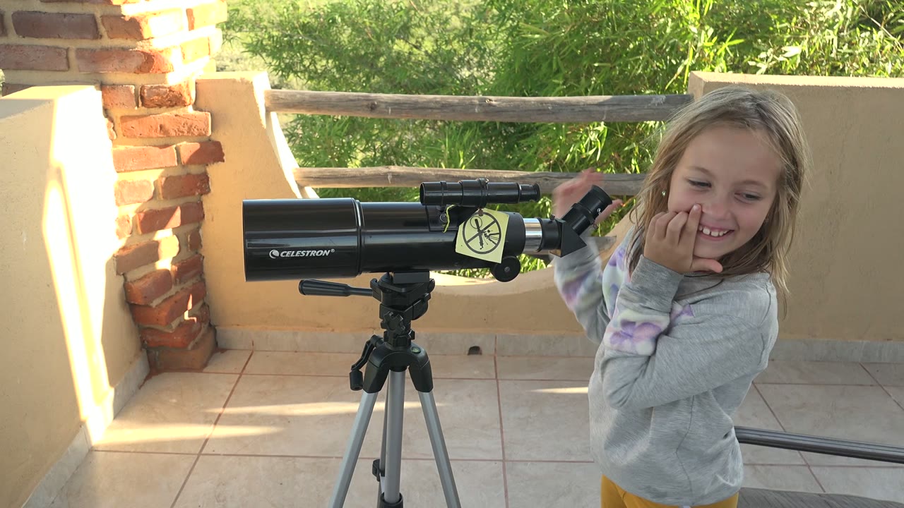 A young girl in San Carlos, Mexico smiles near the right side of the frame as the black telescope and tripod remain centered on the tiled patio.