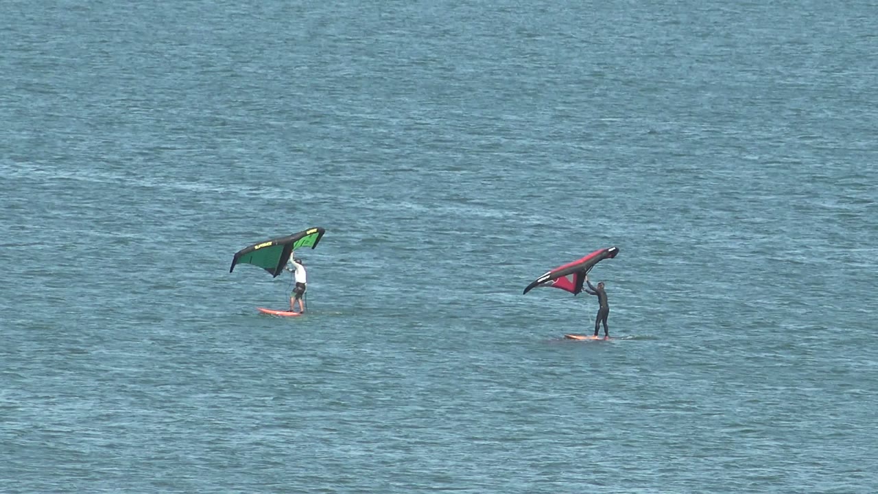 Two distant kite surfers ride on calm ocean water in San Juanico, Baja California Sur, with a green kite on the left and a red kite on the right.