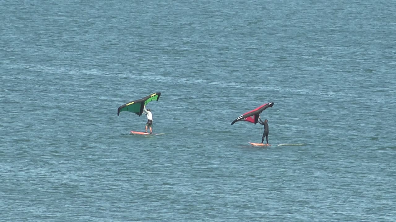Two San Juanico kite surfers hold a green kite and a red kite above their boards while the surrounding ocean surface stays lightly textured.
