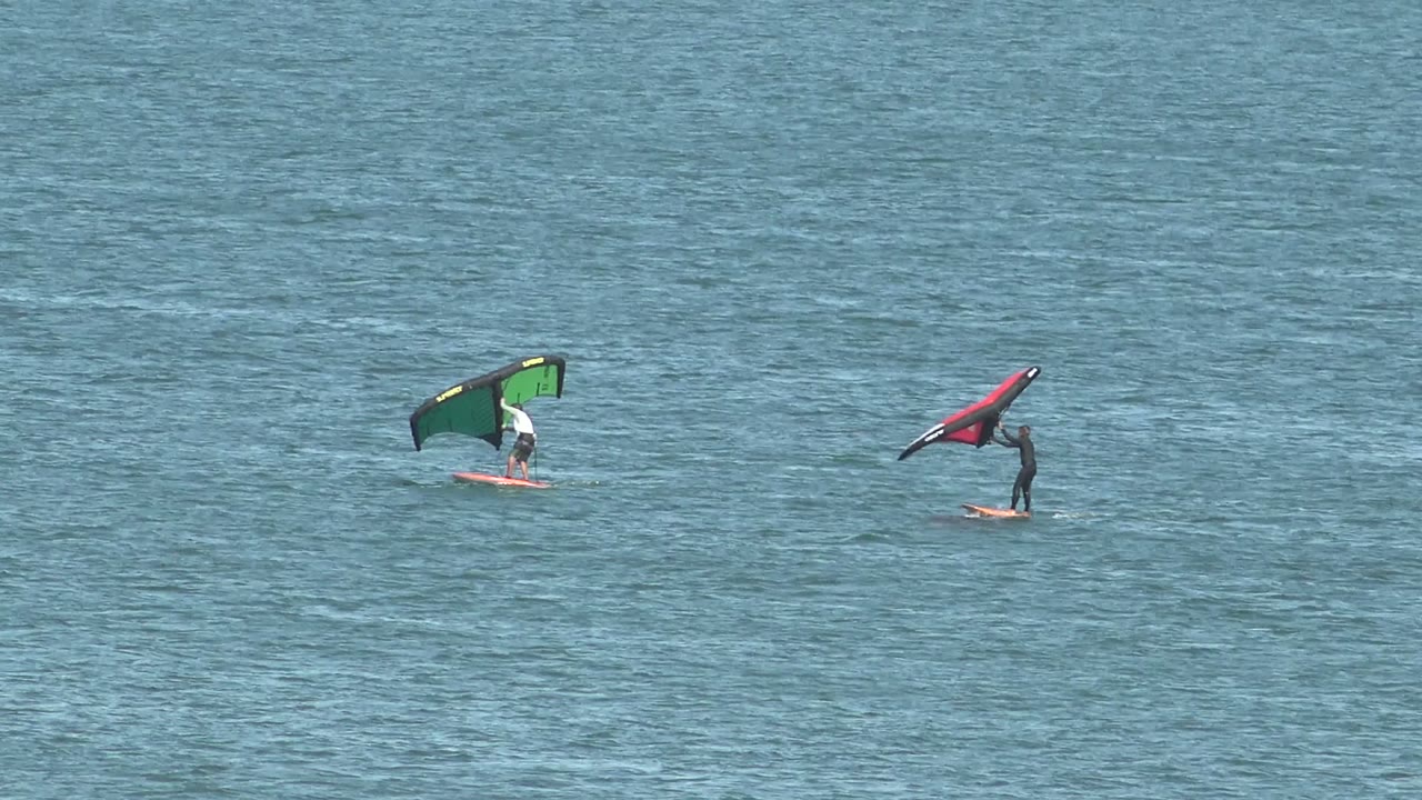 Two distant riders remain visible on open blue water in San Juanico, with the green wing upright and the red wing angled close to the water surface.