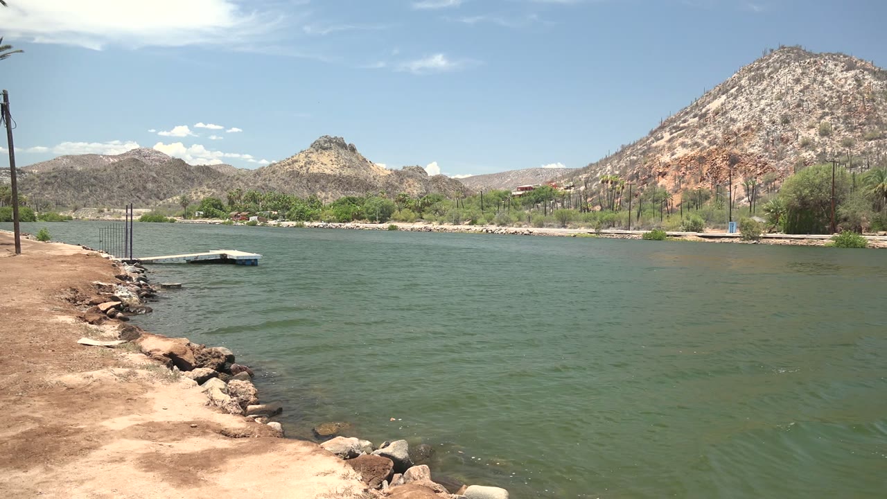 Opening view of green rippling water in Mulege, Baja California Sur, with a rocky dirt bank at left, distant dry mountains, trees, and no visible people.