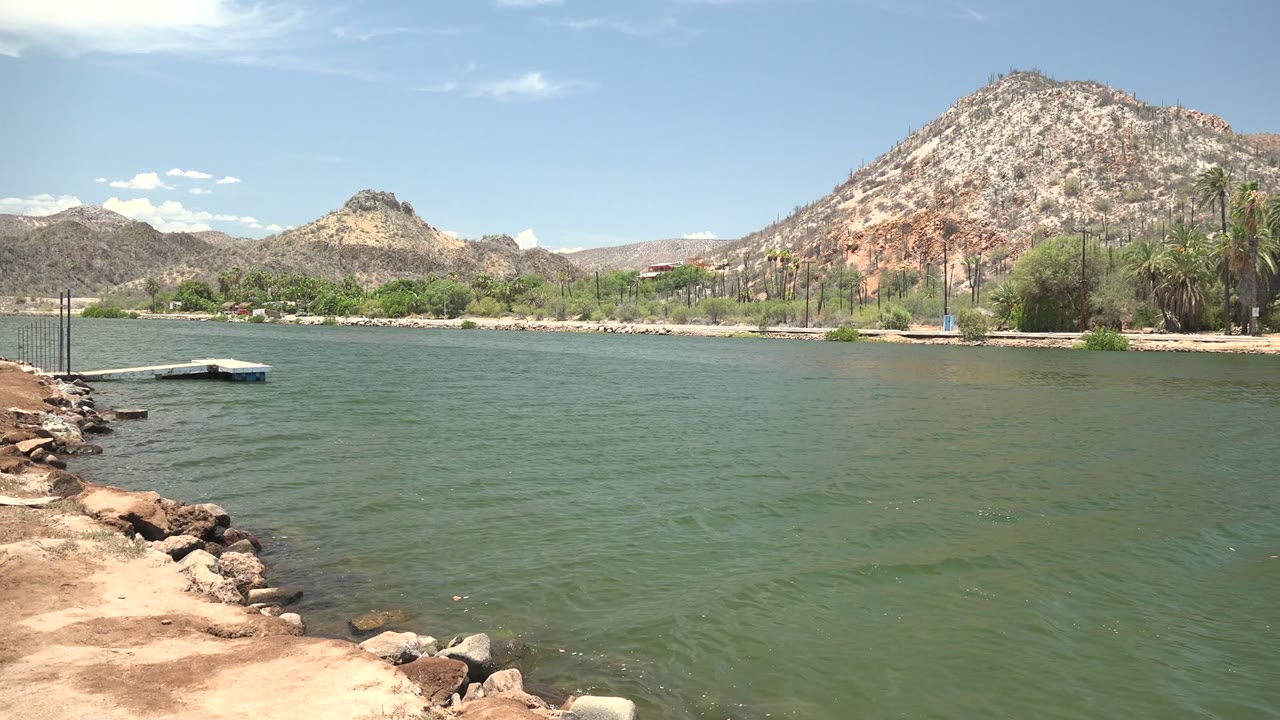 The pan continues across Mulege water, with the rocky bank still at left, a small dock near the far shore, dry hills across the background, and no visible people.