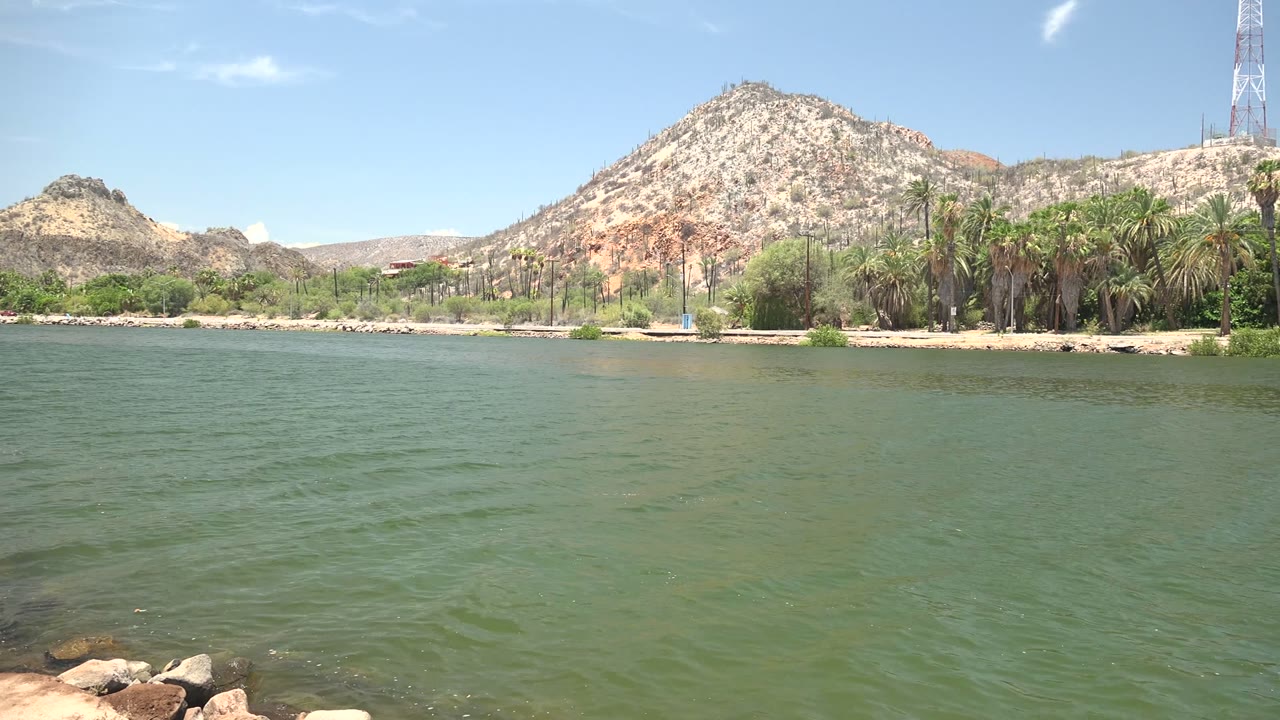 At 0:00 in Mulege, Baja California Sur, green lake water fills the foreground below palms, a rocky hill, a tower, and blue sky, with no visible people.