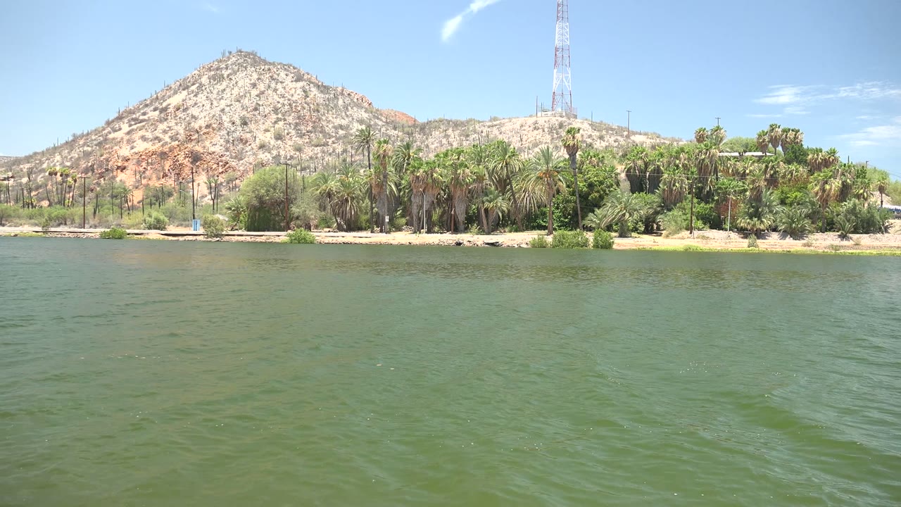Green water ripples below a rocky hill, trees, and tower.