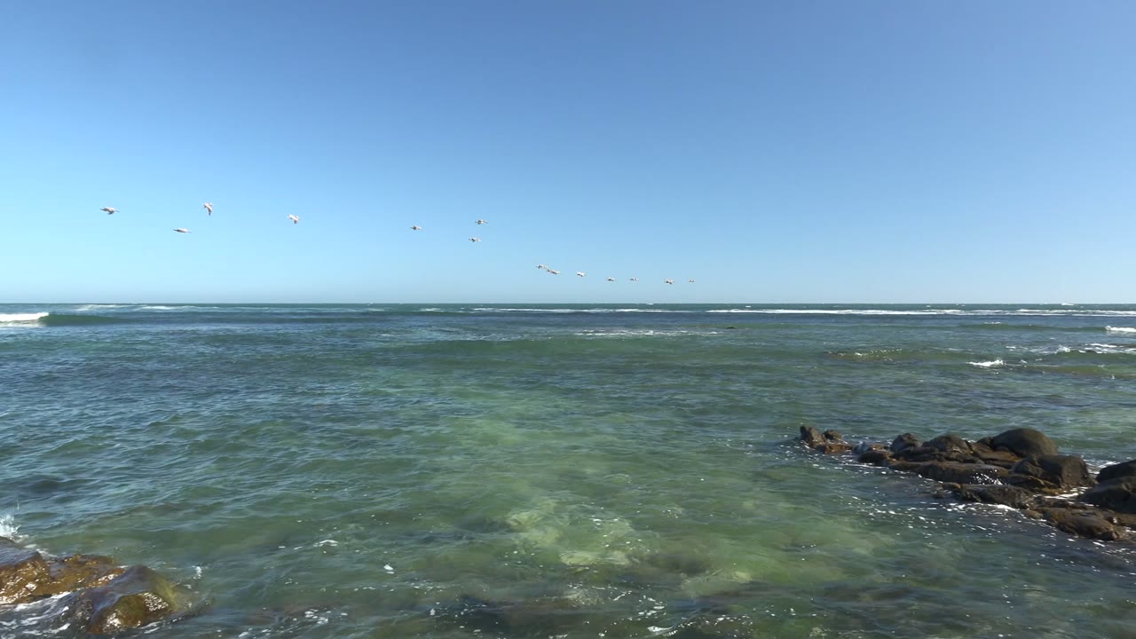 A broad La Bocana seascape shows small waves breaking behind dark rock outcrops, pale green water patches in the foreground, birds near the horizon, and no visible people.
