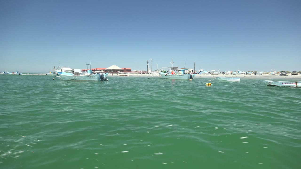 Wide water-level view in La Bocana, Baja California Sur, with green rippled ocean in front, small boats near the sandy shore, distant beach structures, and no visible people.