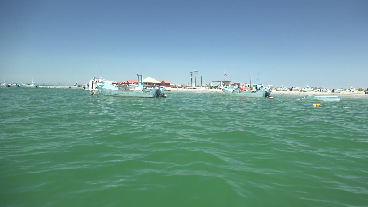 Green water stretches to several small boats along the La Bocana shore, with a pale sandy strip, low structures, and a clear blue sky, and no visible people.