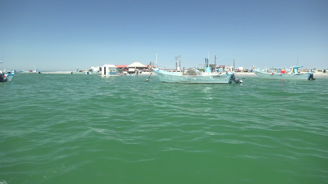 Low view across green rippled water toward anchored fishing boats and shoreline buildings in La Bocana, Baja California Sur, with no visible people.