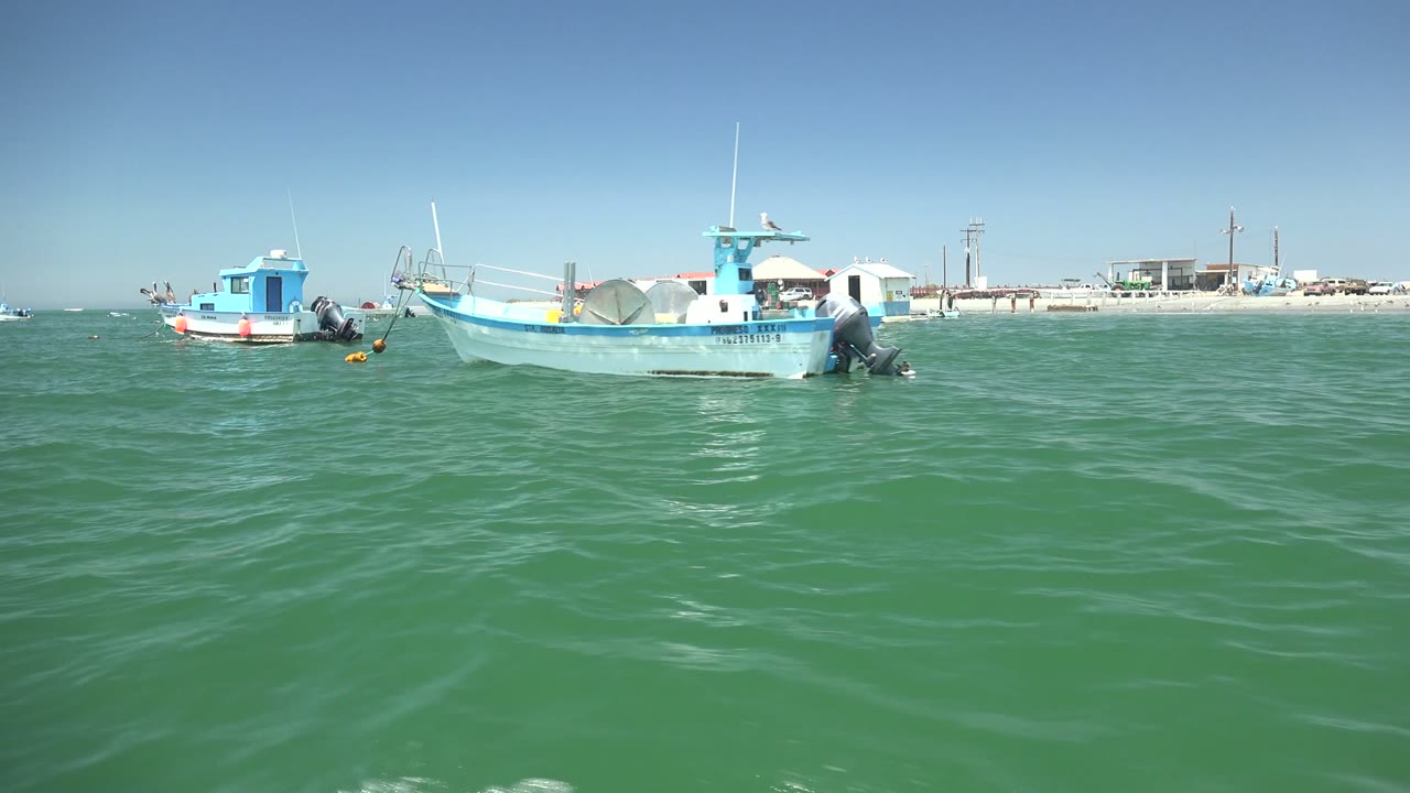 A blue-cabin fishing boat sits closer to the right side of frame in La Bocana, Baja California Sur, with another small boat to the left and no visible people.