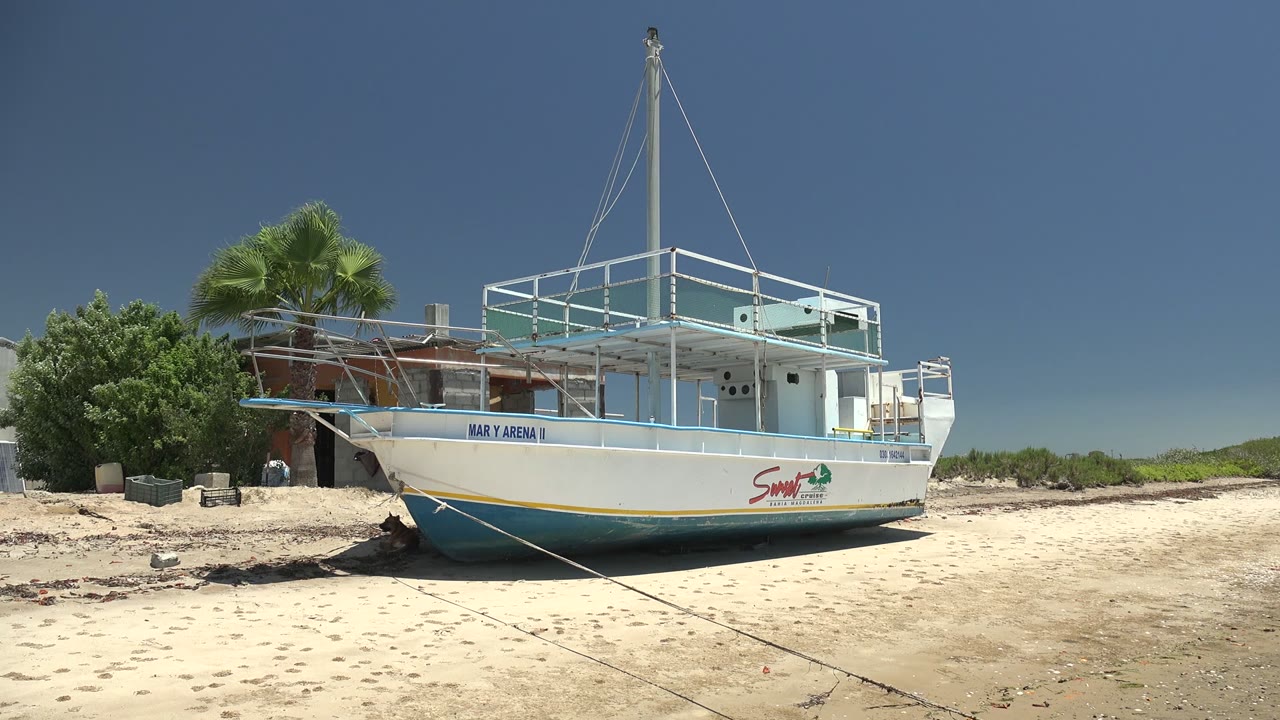 At 0:00 in San Carlos, Baja California Sur, a white boat with blue trim rests on sand, with a rope leading across the foreground, a palm and low building behind it, and a small distant person near the background.