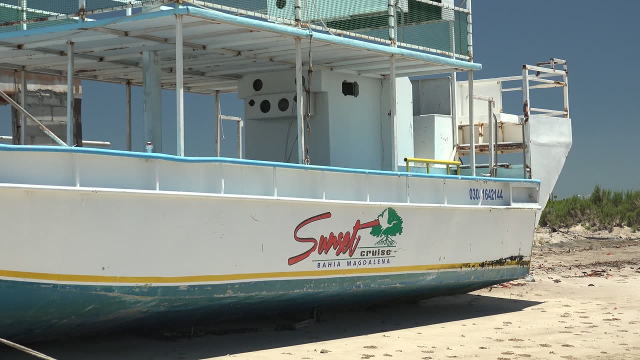 Wide close view of a white and blue boat on a sandy beach in San Carlos, Baja California Sur, with Sunset Cruise lettering on the hull and no visible people.