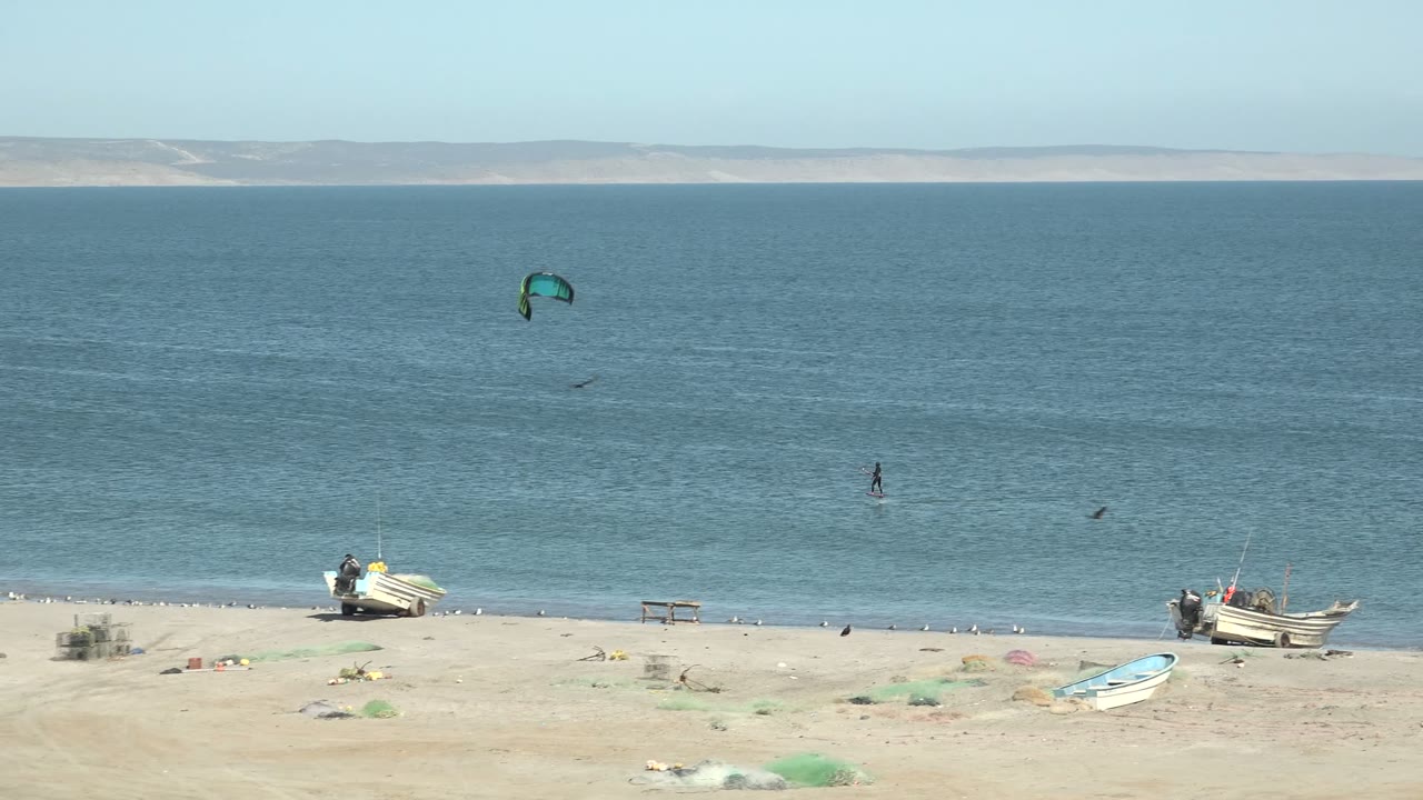 Wide San Juanico, Baja California Sur beach view with a distant kite surfer under a green kite, small boats on pale sand, and low land across the horizon.