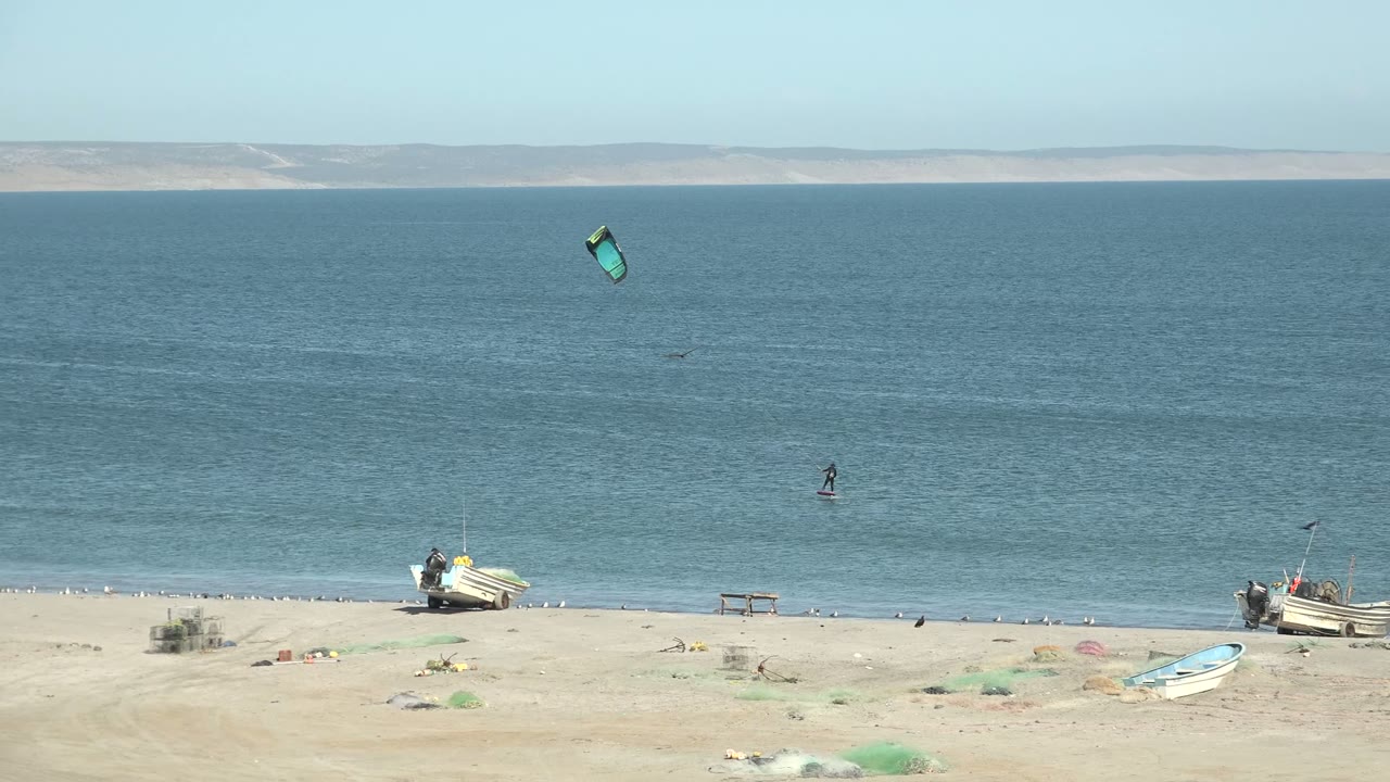 The green kite sits higher over the blue water as the distant kite surfer continues near the sandy San Juanico shoreline with boats and netting below.