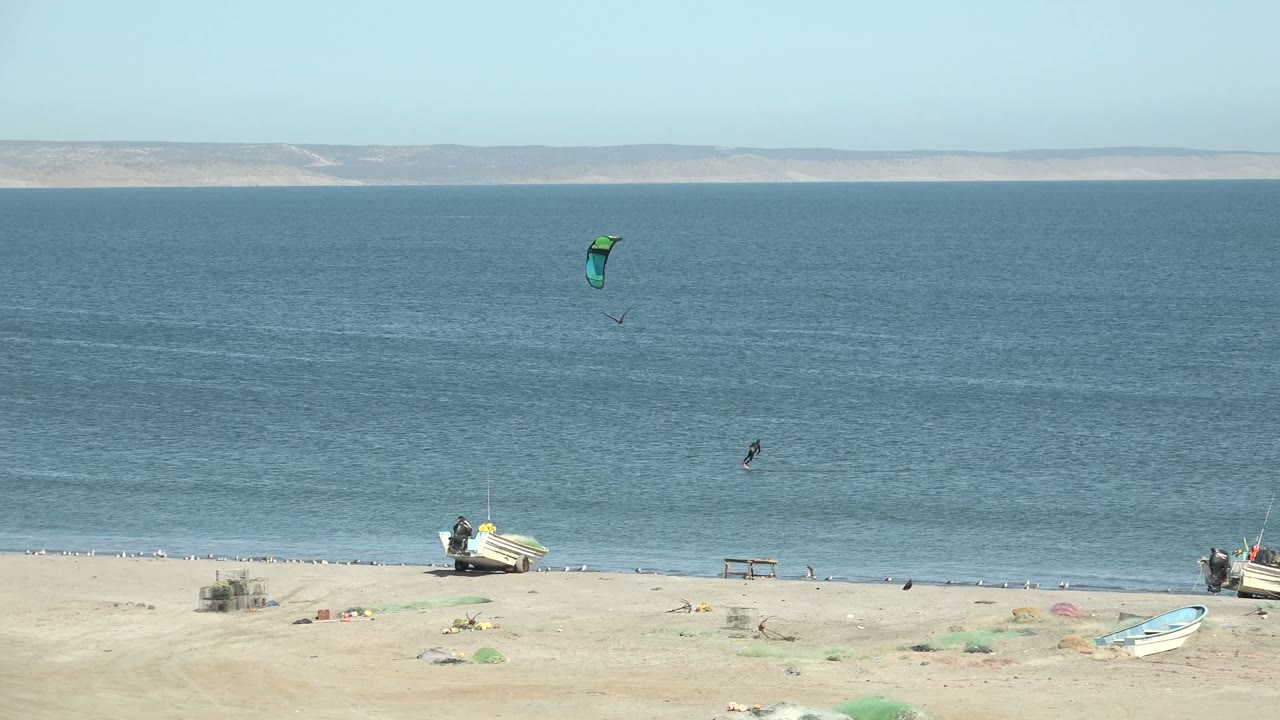 A distant kite surfer moves across the calm ocean near San Juanico while a green kite hangs above and beach boats sit along the lower edge of frame.