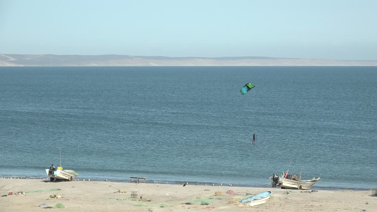 Wide San Juanico beach frame with a green kite above calm blue water, a small kitesurfer offshore, beached boats on the sand, and distant land across the horizon.