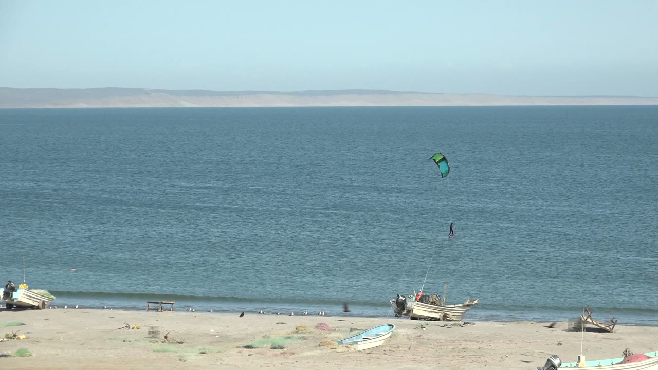 Wide San Juanico frame with the green kite near the upper right of the water, the small rider below, gentle surf at the beach, and beached fishing boats in the foreground.