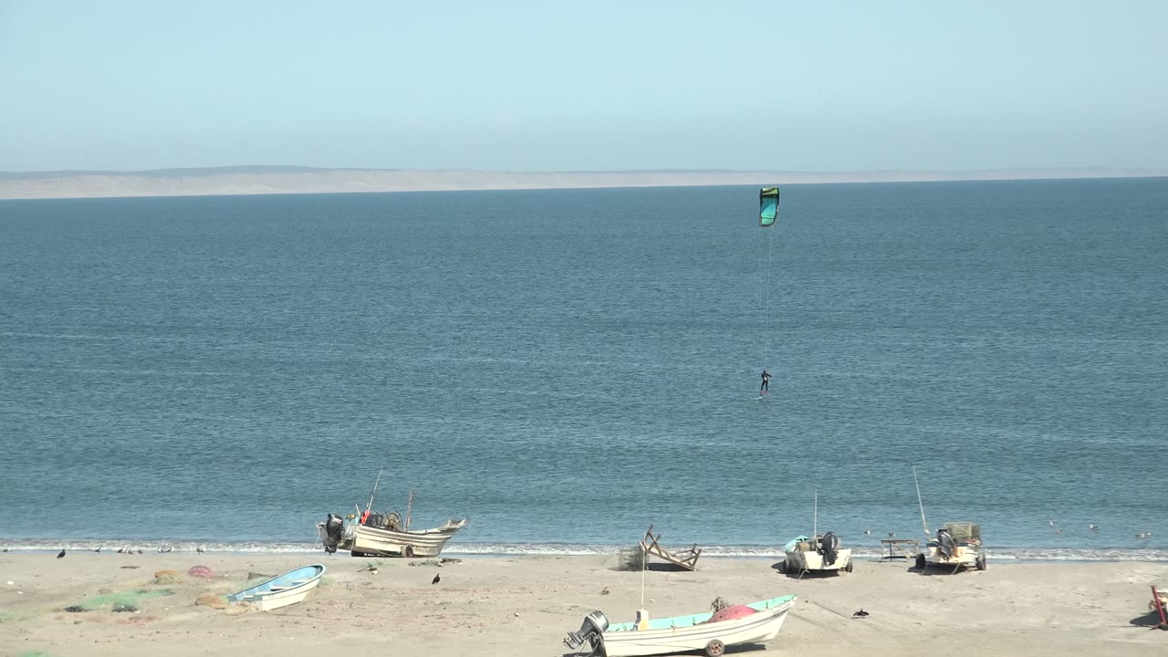 San Juanico, Baja California Sur beach with small fishing boats on pale sand, blue ocean beyond, and tiny kite surfers offshore near the start of the clip.