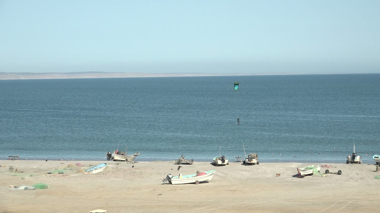 San Juanico shoreline with multiple fishing boats resting on the beach, low waves at the water's edge, and a small kite and rider visible against the blue ocean.