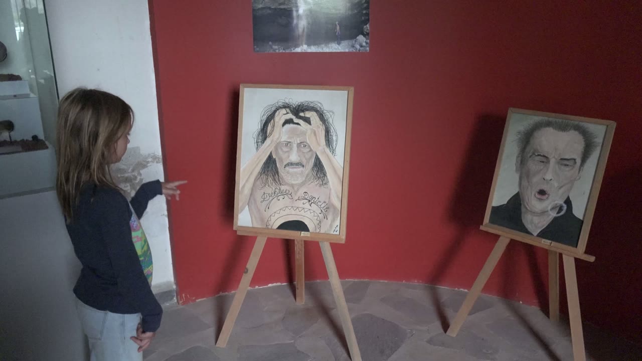 A girl in profile points toward two framed artworks on wooden easels inside a red-walled gallery room in Mulege, Baja California Sur.