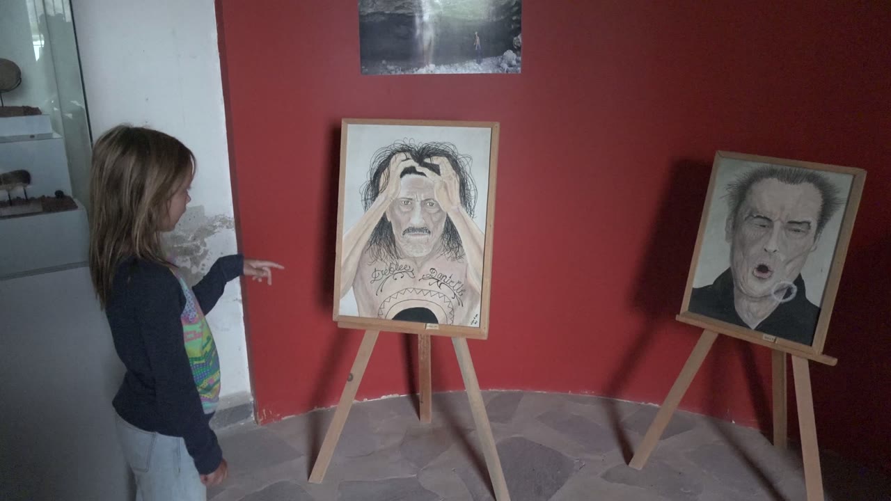 Girl stands at left pointing near two easel-mounted artworks.