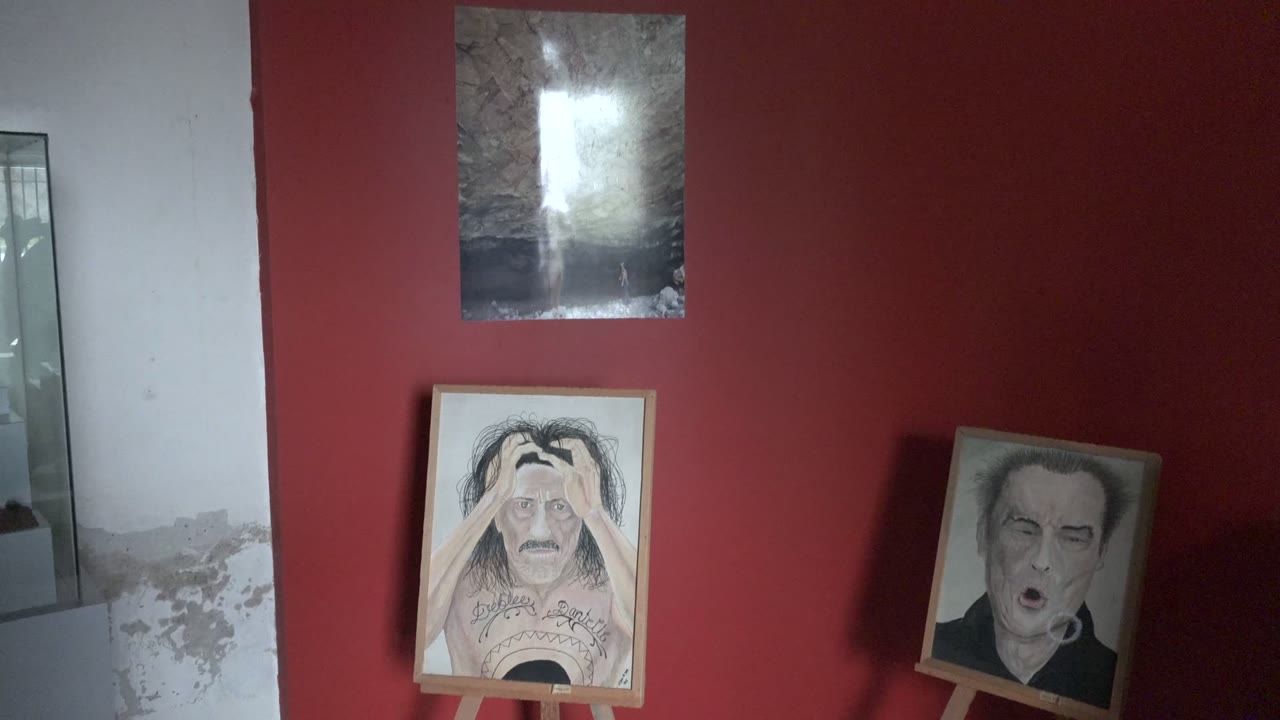 Opening frame of a Mulege indoor art exhibition with two portrait paintings on wooden easels, a red wall, a dark reflective artwork above, a mirror at left, and no visible people.