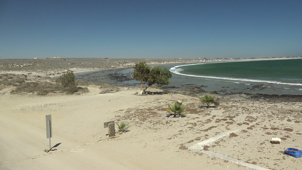 Opening frame of La Bocana beach in Baja California Sur, Mexico with pale sand, a signpost at left, low plants, a small tree near the shore, green-blue water, clear sky, and no visible people.