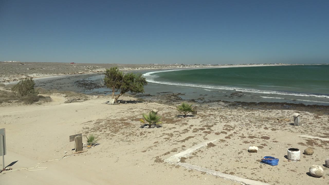 La Bocana coastal frame with the signpost and foreground beach objects shifting toward the right side, a blue container on sand, sparse vegetation, rolling waves, and no visible people.