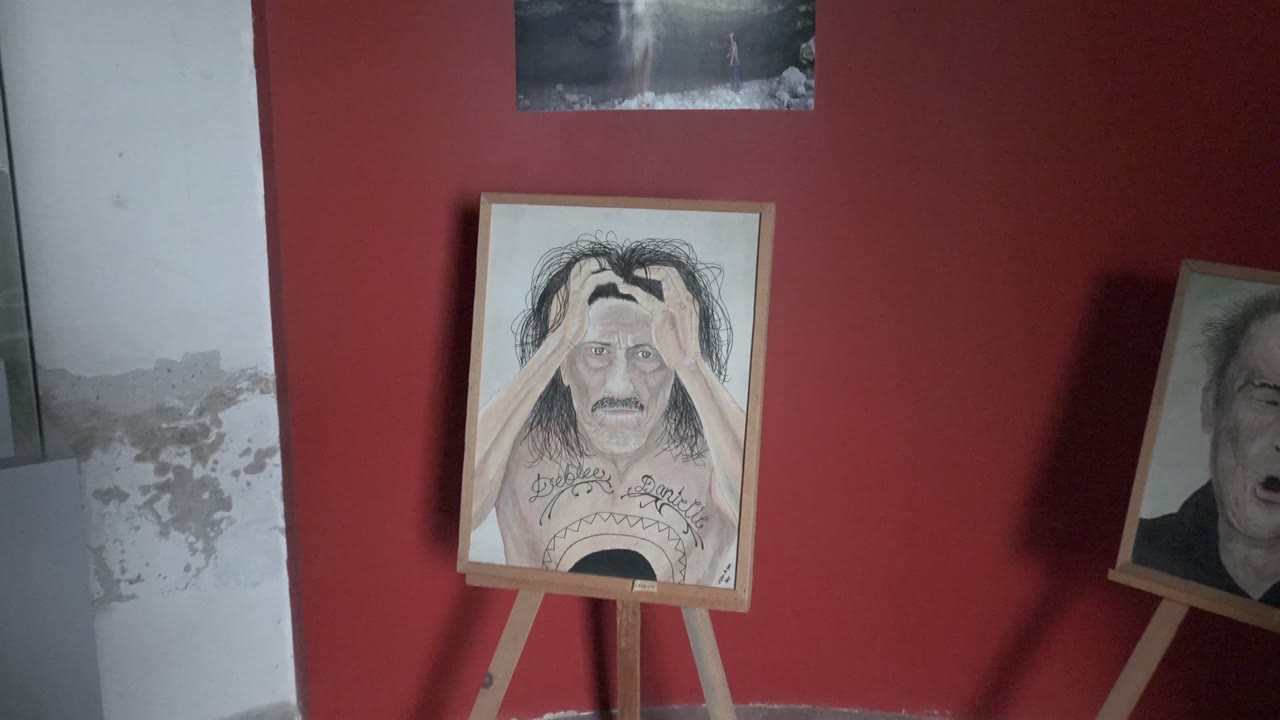 Wide Mulege art-room view with a framed portrait painting on a wooden easel, red wall behind it, white peeling wall at left, a second easel at right, and no visible people.