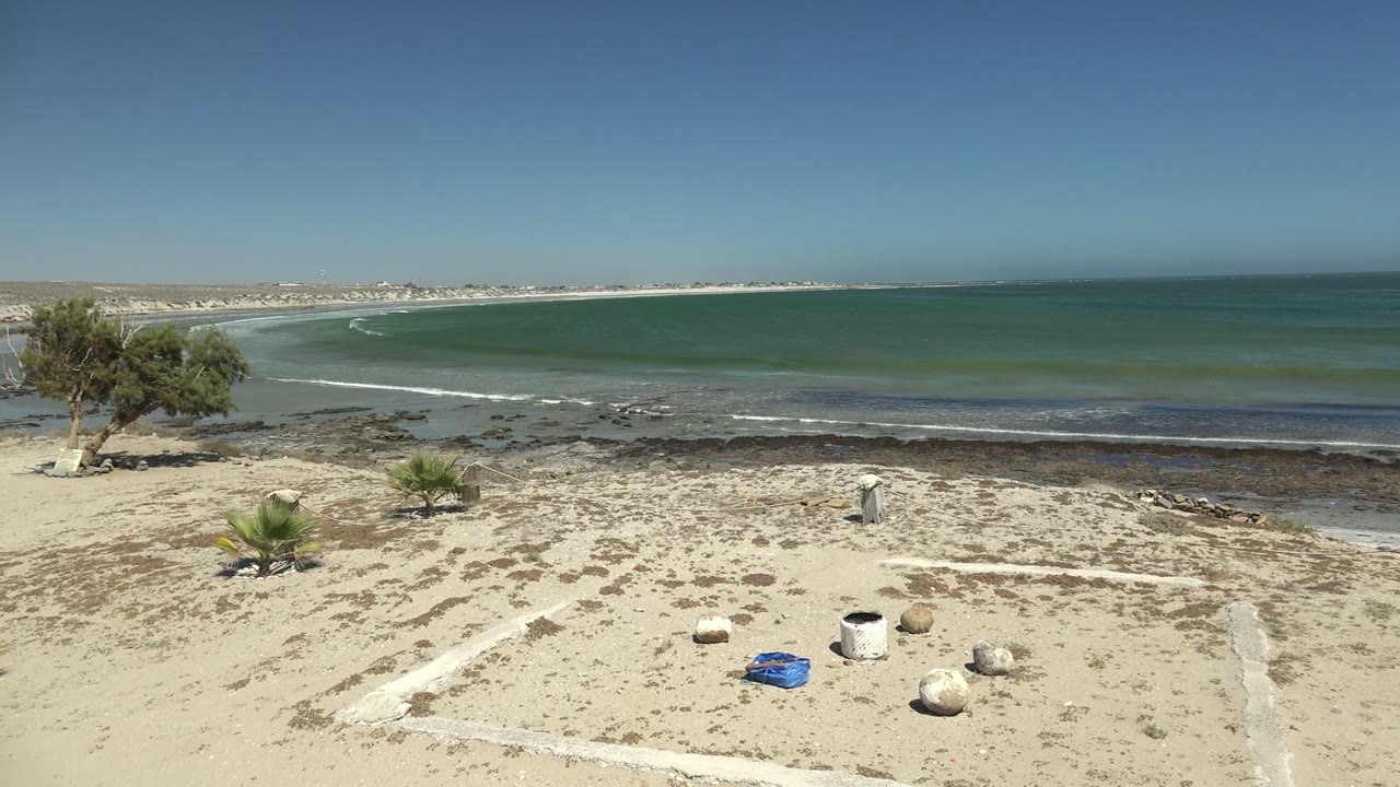 Opening view of La Bocana beach with sandy foreground, small palms, scattered rocks, distant surf, green-blue ocean, clear sky, and no visible people.