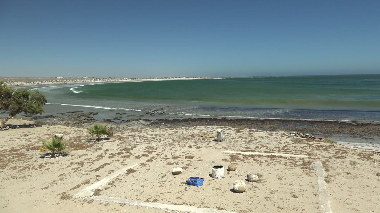 Beach foreground with white sand lines, palms, rocks, and ocean surf beyond.