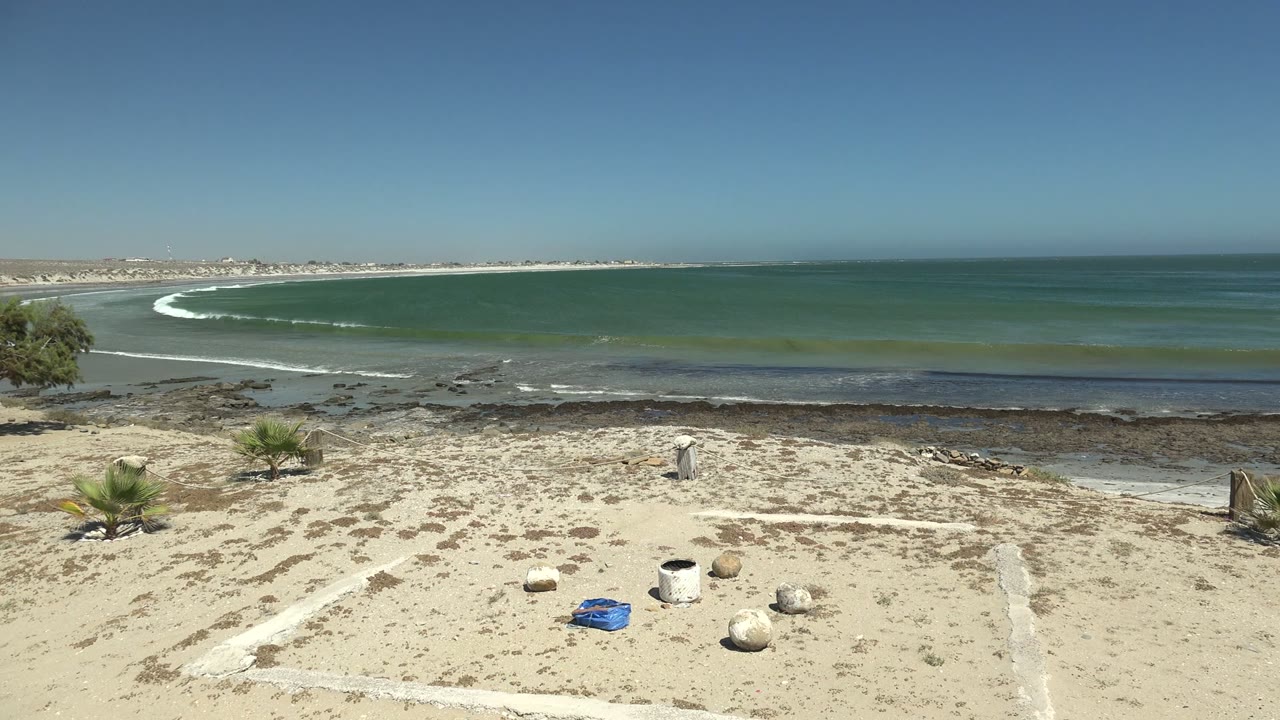La Bocana beach frame with a bright curved wave offshore, sandy foreground markings, small palms, scattered rocks, blue-green water, and no visible people.