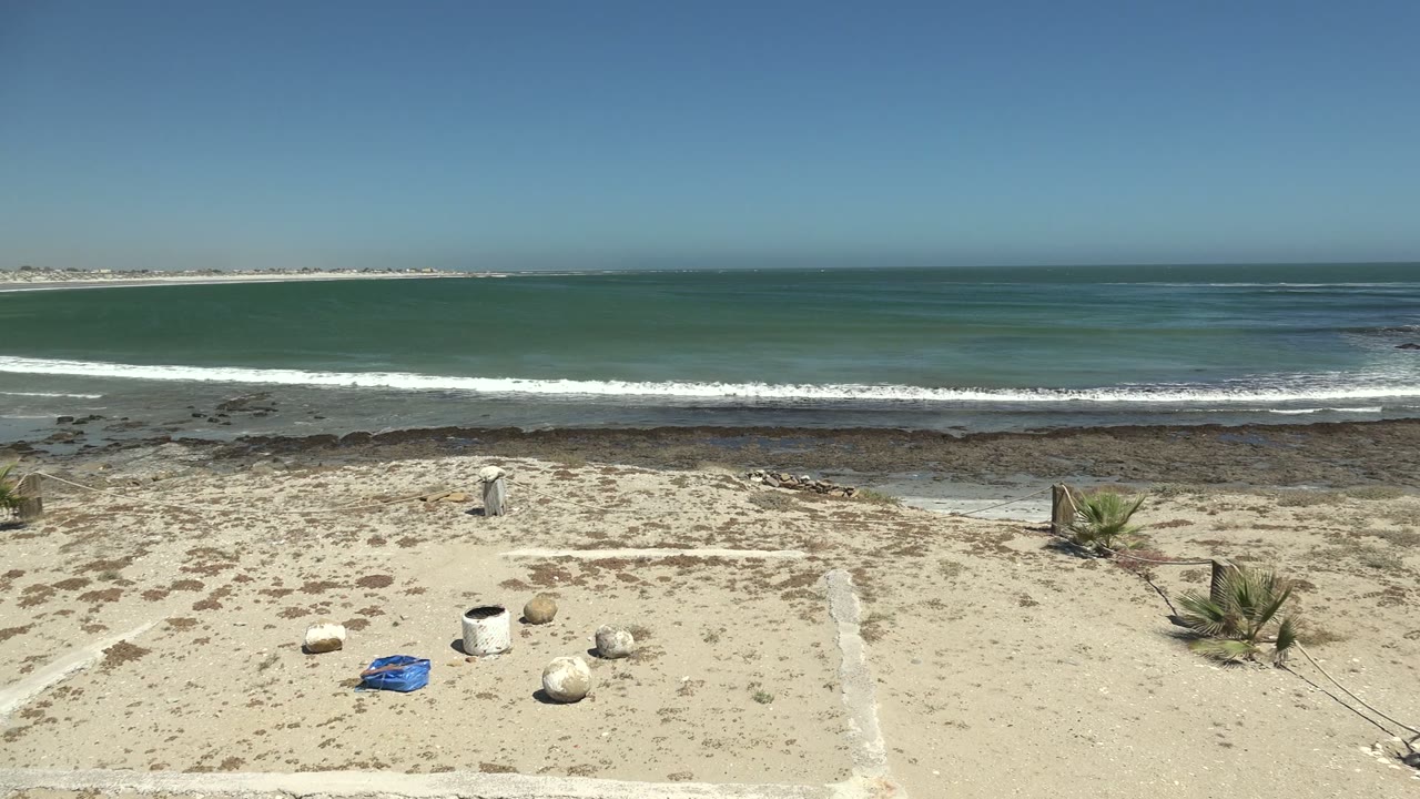 Wide La Bocana beach view with pale sand, scattered rocks, a white bucket, a blue object, low waves over a rocky shore, green-blue ocean, clear sky, and no visible people.