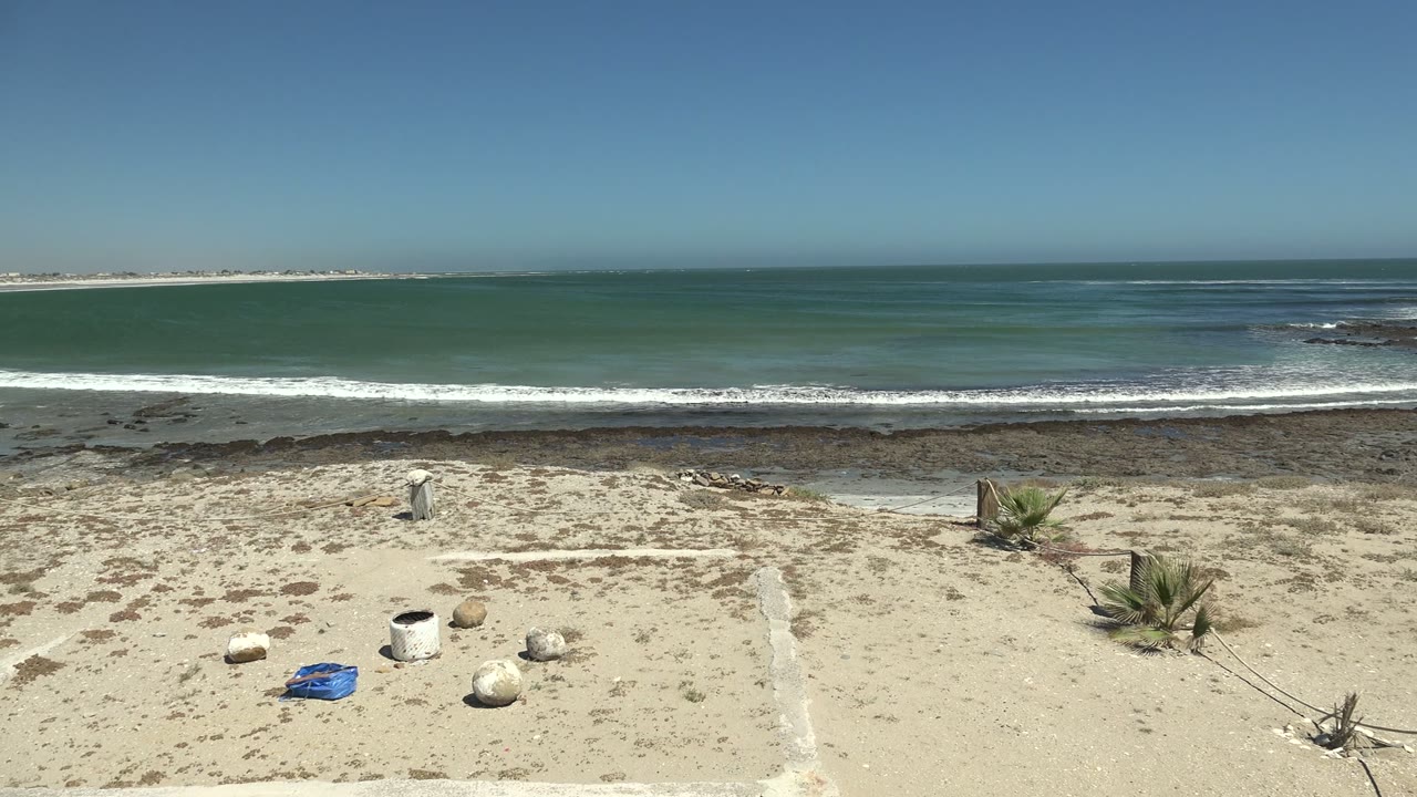 La Bocana shoreline frame with the blue object and white bucket still on the sand, small plants near center right, white surf bands, a dark rock shelf, and no visible people.
