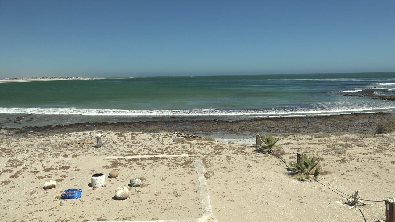 La Bocana beach view during the pan with foreground rocks and a blue object lower left, small desert plants near the sand path, white waves breaking on the rocky shoreline, and no visible people.
