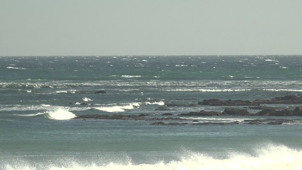 Wide view of choppy blue-green ocean at La Bocana, Baja California Sur, with white surf hitting dark rock shelves and no visible people.