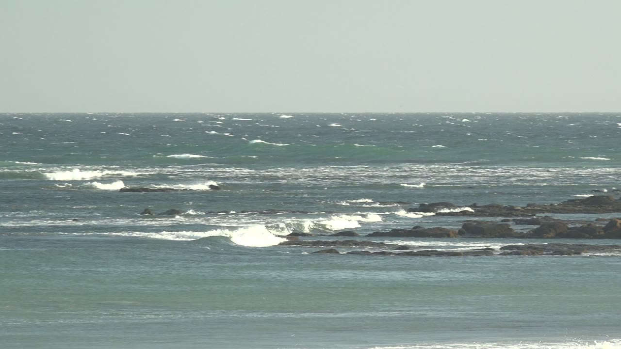 A wide La Bocana shoreline frame shows shallow green water in front, dark rocky reef in the middle, and breaking white waves farther out with no visible people.