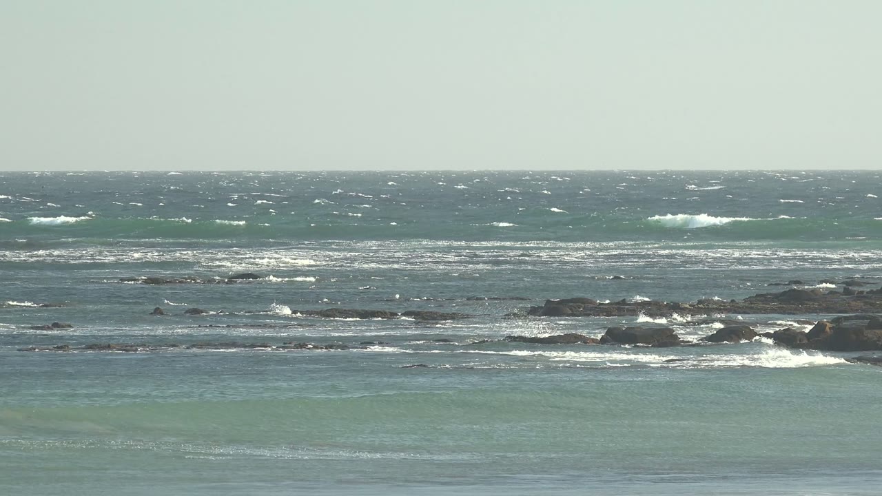 Wide view of blue-green ocean waves and whitecaps breaking over dark rocks at La Bocana, Baja California Sur, with pale sky above and no visible people.