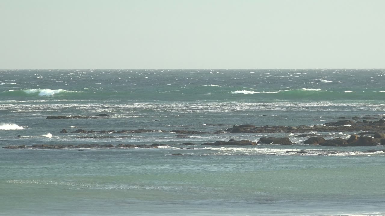 A broad band of wind-ruffled ocean at La Bocana shows scattered whitecaps, exposed dark rocks, pale green nearshore water, and no visible people.