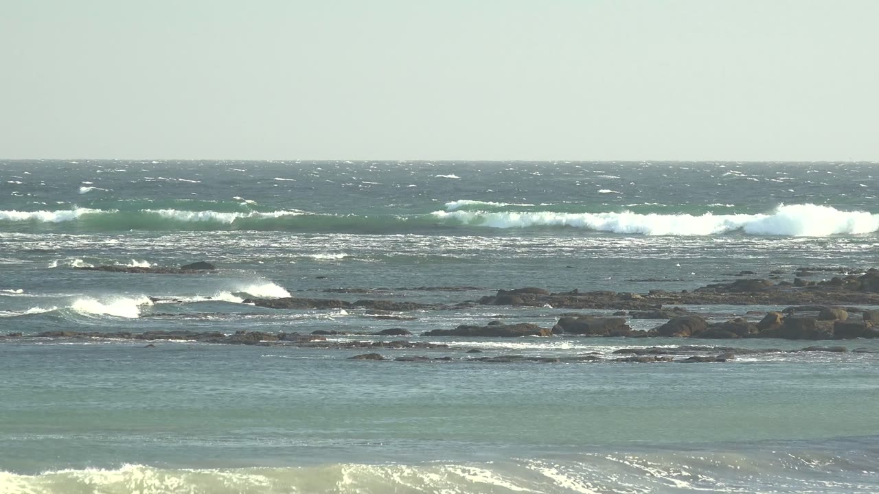 Wide view of La Bocana ocean surf with blue-green water in front, low rocks across the middle, white waves behind them, and no visible people.