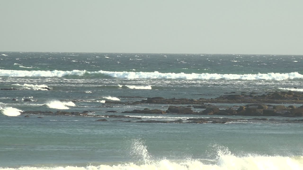 A burst of white wave spray rises near the right side of the La Bocana rocky shoreline, with layered surf and no visible people.