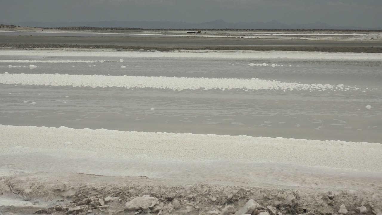 Wide view of a Guerrero Negro salt flat with shallow gray water, white salt deposits, a rough rocky foreground, distant hills, and no visible people.
