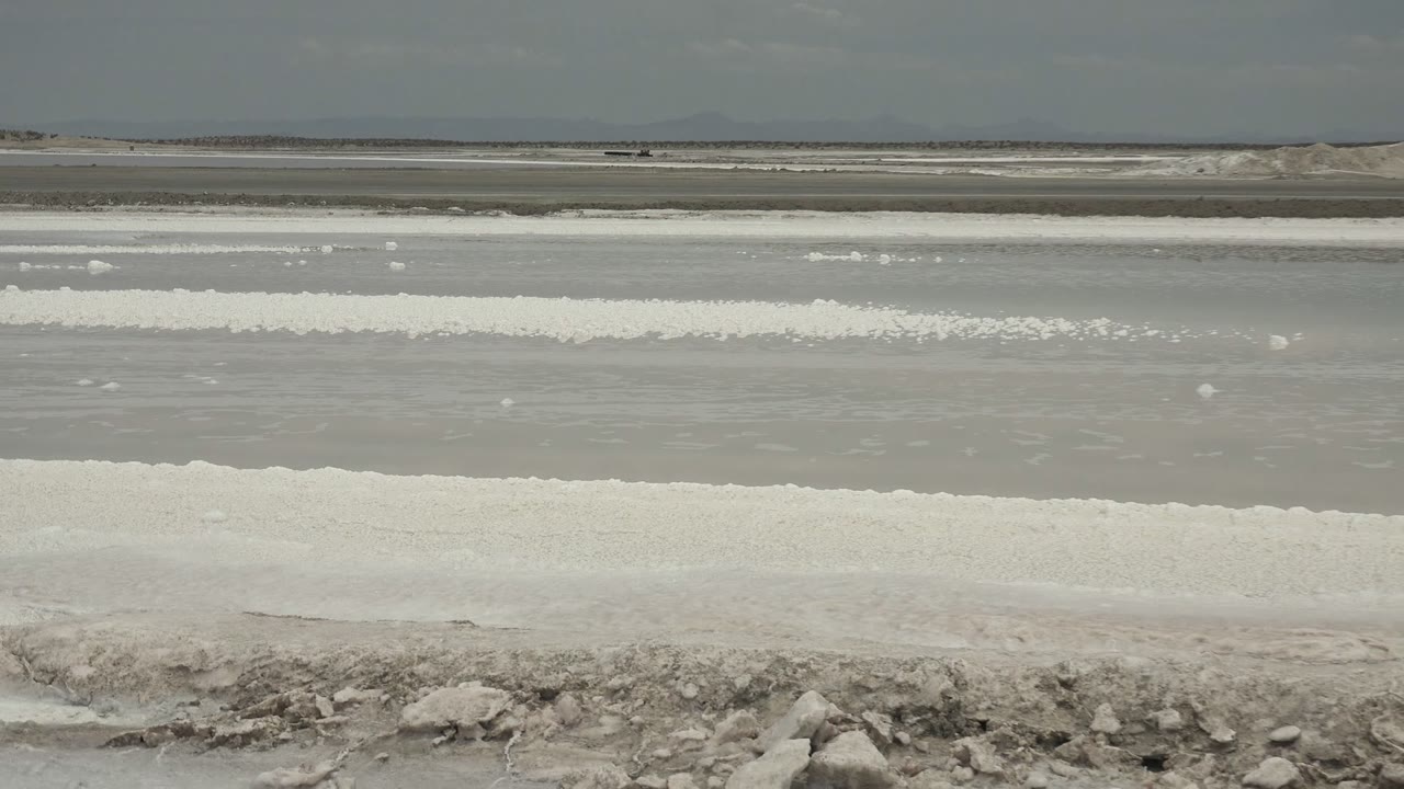 Salt flat water and white mineral ridges under a brighter cloudy sky.