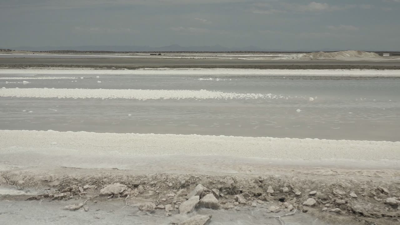 Layered Guerrero Negro salt flat scene with gray shallow water, thick white salt bands, foreground rocks, distant low hills, and no visible people.