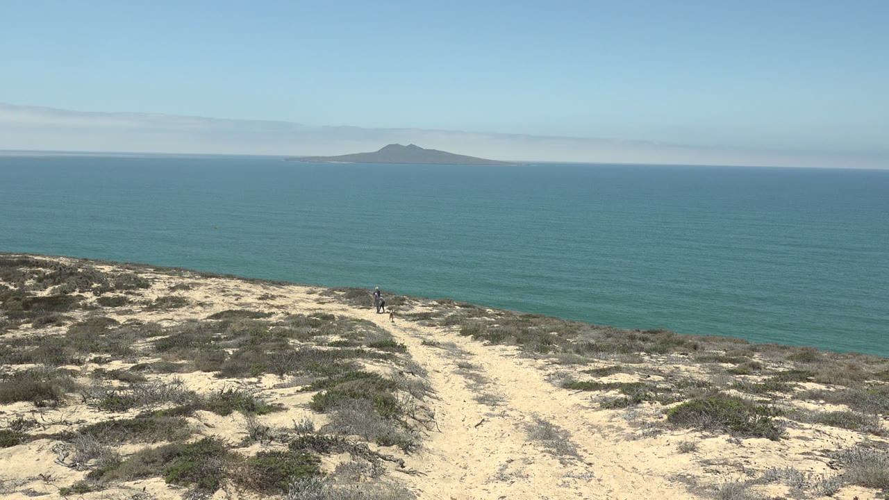 San Quintin, Mexico coastal hillside with pale sand and scrub in the foreground, the ocean stretching across the frame, a distant island near the horizon, and a small person still visible near the trail.