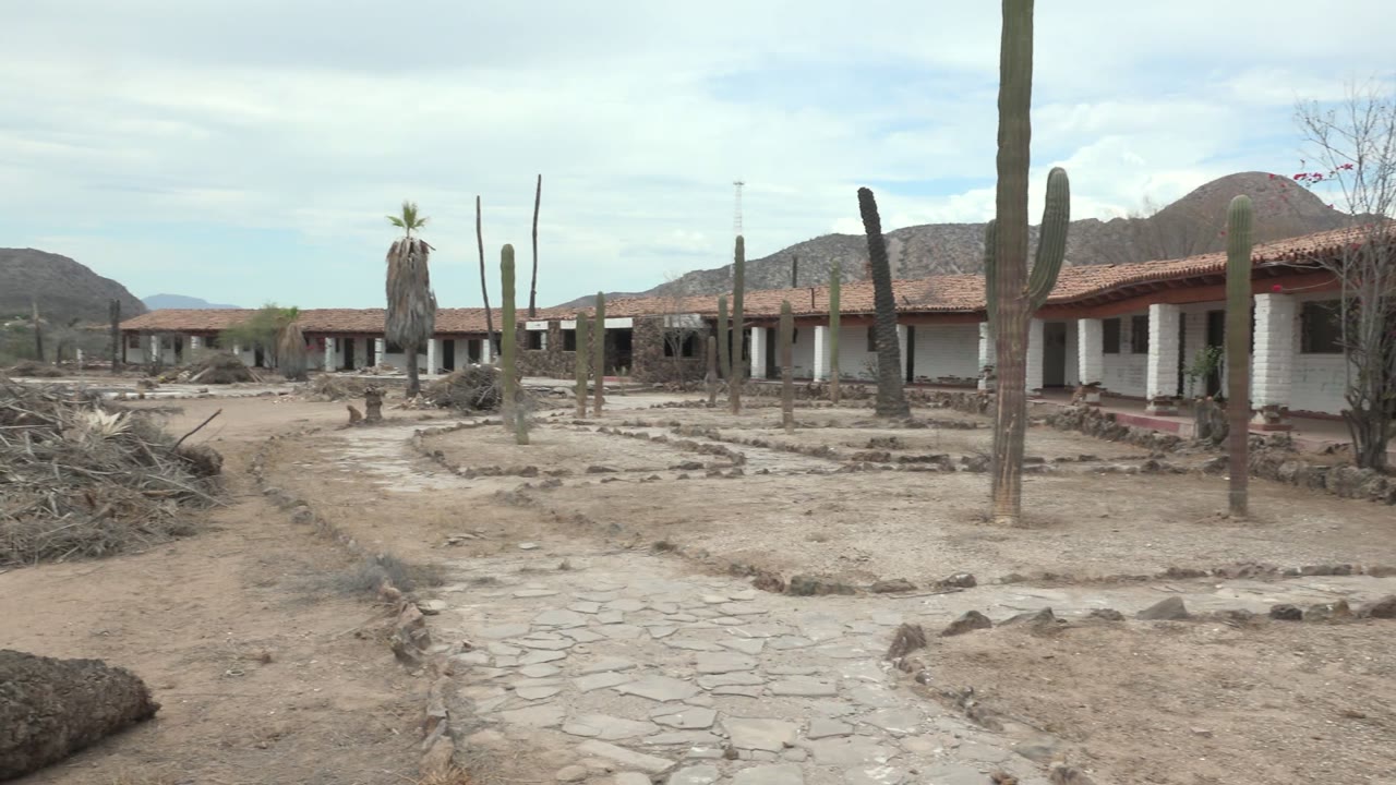 Opening frame of a Mulege desert homestead with tall cacti, sandy ground, a stone path, low red-roofed buildings, distant mountains, and no visible people.